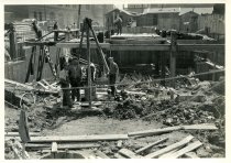 Digital scan of a sepia-toned photo of three workers posing around a bucket suspended from a tripod in front of the foundation for the ventilation building on the construction site for the Detroit terminal of the Detroit Windsor Tunnel, as taken from the 