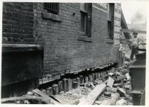 Digital scan of a sepia-toned photo taken of the underpinning put in place beneath the north wall of a brick building at 216 Randolph Street during the construction of the Detroit-Windsor Tunnel