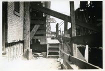 Digital scan of a sepia-toned photo of steps leading to an elevated walk way that runs along the underpinning of the buildings on the east side of Randolph Street between Henry Street and Atwater Street facing south, taken during the construction of the D