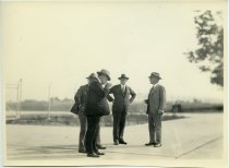 Black and white photographic print of four men gathered in discussion, smoking cigarettes at the Packard Proving Grounds