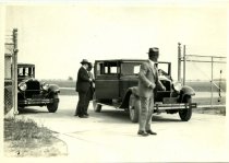 Black and White photographic print of three men standing near two Packard automobiles at a chain-link fence gate