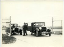 Black and White photographic print of a man helping another man into one of two Packard automobiles at a chain-link fence gate