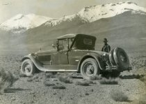 Black and white photographic print depicting a man standing behind a Packard automobile in a desert