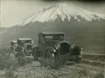 Black and white photographic print depicting a line of three Packard automobiles with a snowcapped mountain in the background