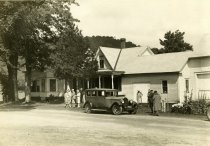 Black and white photographic print of a Packard sedan parked in front of a house and surrounded by a group of people in Plymouth Notch, VT