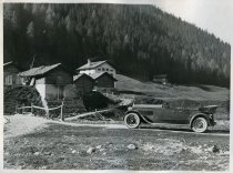 Black and white photographic print of a left side profile view of a Packard 8 convertible parked on a road near several houses in the Swiss Alps, with a wooded mountain in the background