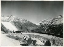 Black and white photographic print showing a left side profile view of a Packard 8 convertible parked along the side of a mountain road in the Swiss Alps, with a man standing to the left of the Packard and mountains in the background