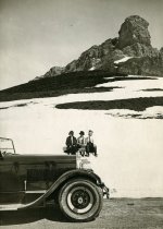 Black and white photographic print featuring a right side profile view of the front half of a Packard 8 convertible, parked along the side of a road in the Swiss Alps