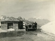Black and white photographic print showing a front end view of a Packard 8 convertible which is parked on a mountain road, near a stone building, in the Swiss Alps