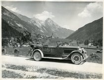 Black and white photographic print showing a right side profile view of a Packard 8 convertible parked along the side of a road that is on a mountain overlooking a town in the Swiss Alps