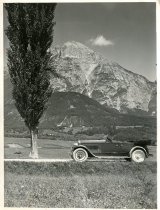Black and white photographic print showing a left side profile view of a Packard 8 convertible parked near a tree on the side of a road in the Swiss Alps