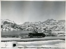 Black and white photographic print of a right side profile view of a Packard 8 convertible parked on a road near a small body of water in the Swiss Alps