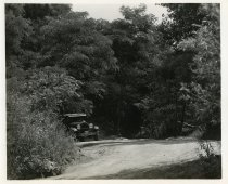 Black and white photographic print taken outside depicting a Packard automobile from the front driving up a hill on a dirt road