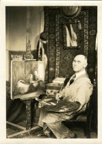 Sepia-toned portrait photo of Charles Waltensperger in his studio posed holding a brush and palette seated in front of a painting of a woman sewing beside a baby