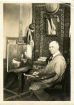 Sepia-toned portrait photo of Charles Waltensperger in his studio posed holding a brush and palette seated in front of a painting of a woman sewing beside a baby
