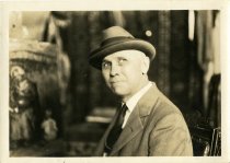 Sepia-toned portrait photo of Charles Waltensperger in his studio posed sitting while clad in a coat, tie, and hat