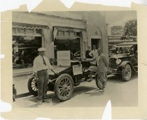 Black and white photographic print taken outside depicting an old Packard automobile with two men standing beside it