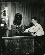 One black and white photograph of a man who is seated at a desk with various pieces of radio equipment