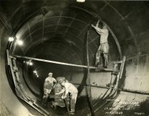 One sepia-toned photograph showing three workmen at work inside a large diameter sewer tunnel