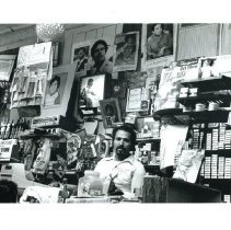 Black & white image of shopkeeper behind a cluttered counter