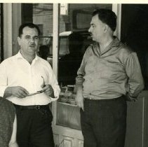 Black & white photo of two men standing outside of shop