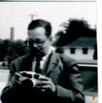 Black and white photograph of man holding camera at his chest