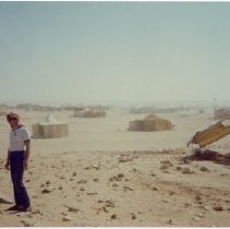 Color photo of two men standing in desert setting w tents behind