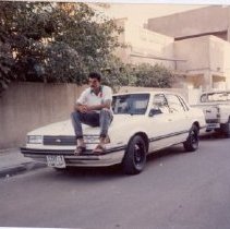 Color photo of man sitting on hood of white car