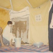 Color photo of three people in a tent, a young boy sits w a bucket