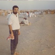 Color photo of man holding red keffiyeh in front of refugee camp