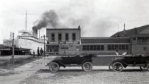 Automobiles on road next to Garage and Supplies, Large boat in background