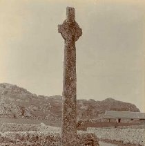 Man with pipe and cap standing at foot of large stone cross near a cottage, at McLean's Cross, Iona, photograph, ca. 1900.