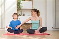 An adult and a child practicing yoga while sitting next to each other. The adult's hand is touching the child's stomach.