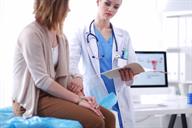 A health care provider holding a folder and speaking with a person in an exam room.