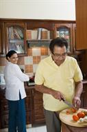 Two adults cooking together. One is at the stove while the other slices tomatoes.