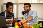 Two adults cooking together in a kitchen. Fruit and vegetables are on the counter in front of them.