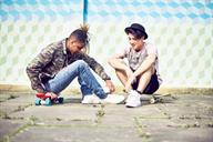 Two teens talking outdoors. One teen is sitting on a skateboard.