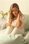 A person sitting on a bed and checking their temperature with a thermometer. 