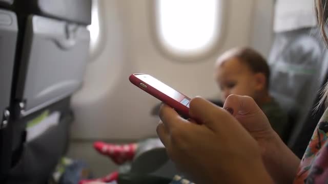 Woman types on cellphone, her baby sits next seat. Travelling family aboard