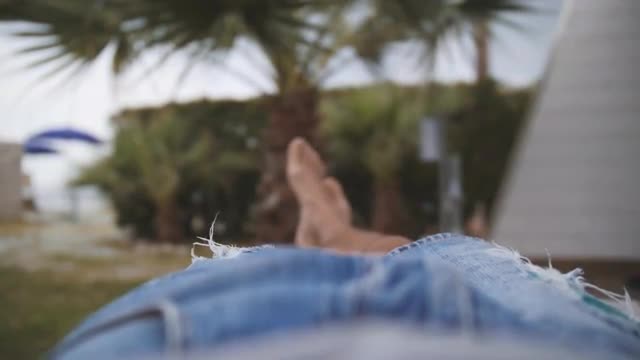 Legs of barefoot man in jeans shorts lying under palm trees. Relaxing guy. POV