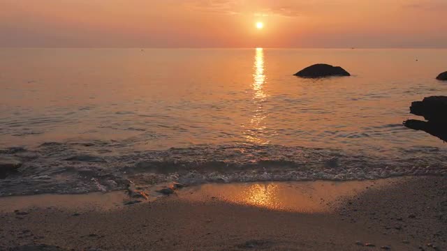 Sun sets into the sea. Water reflects sunlught. Waves roll on sandy beach