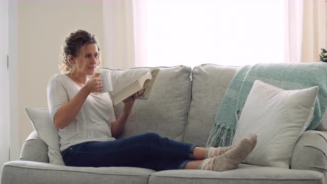 Woman relaxes on the couch while reading a book and enjoying a warm drink 8K