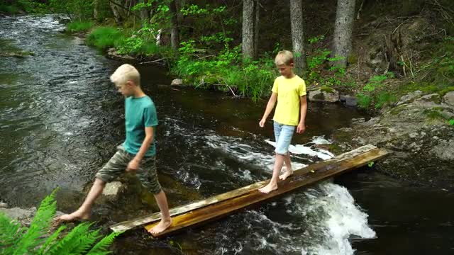 two barefoot boys walking on planks over streaming water in river
