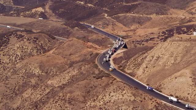 LONG AERIAL traffic with convoy of trucks on highway through desert mountains, California