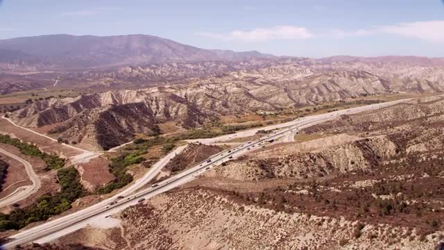 AERIAL POV over desert mountain to LONG AERIAL traffic on highway, California