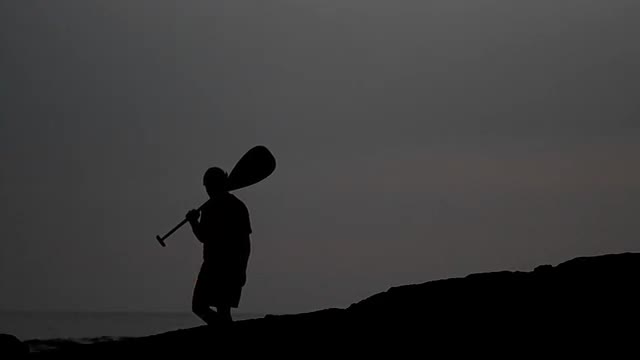 Native Hawaiian walks in silhouette holding a paddle.