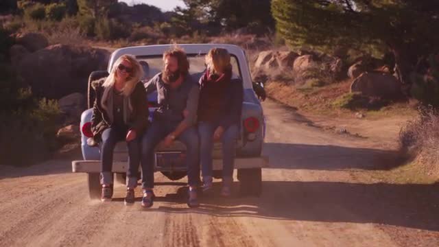 A group of friends sit in the back of a pickup truck as it drives along a rural road.