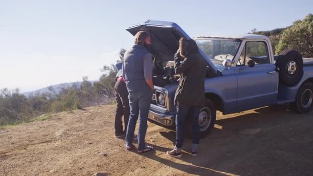 A group of friends stand around an old blue pickup truck with its hood up on the side of a rural roa