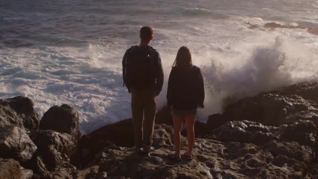 A couple stands on a rocky shore as waves roll in and crash.