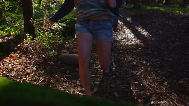 A couple run down a path and leap over a log in a forest.